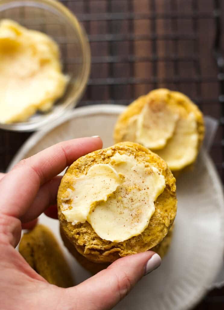 Hand holding a pumpkin biscuit topped with honey butter.
