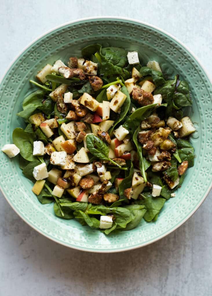 Bowl of fall salad ingredients like baby spinach, chopped apples, brie cheese, and candied pecans.