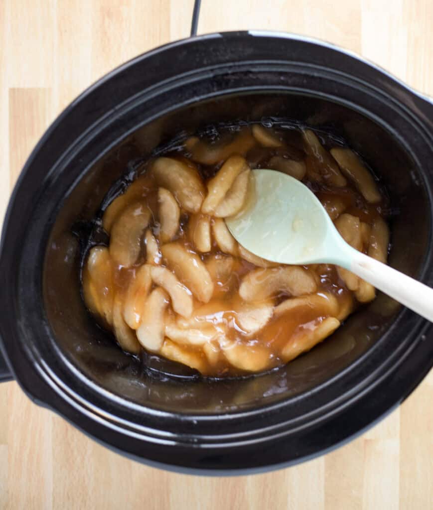 A spatula spreading apple pie filling across the bottom of a slow cooker.