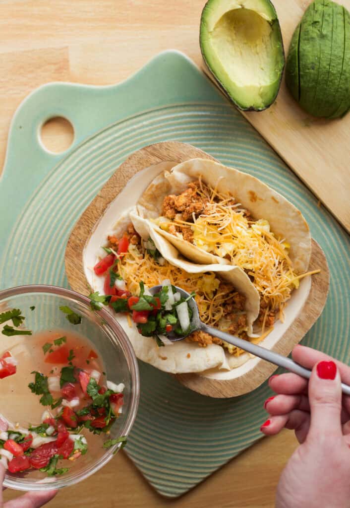 a hand adding toppings to chorizo and egg tacos