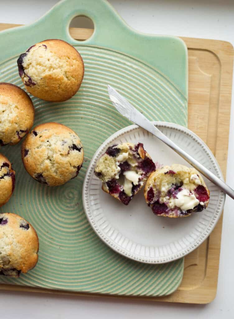 A buttered blueberry muffin on a plate next to a bunch of bakery-style blueberry muffins